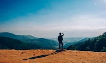 A lone traveler stands on a hilltop, gazing over a vast mountain landscape under a clear blue sky.